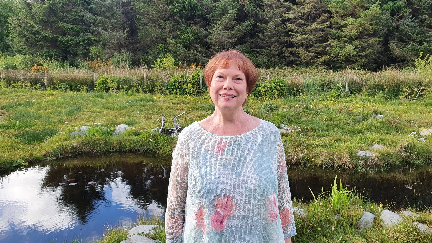 A smiling woman standing in front of a pond set in a green field.