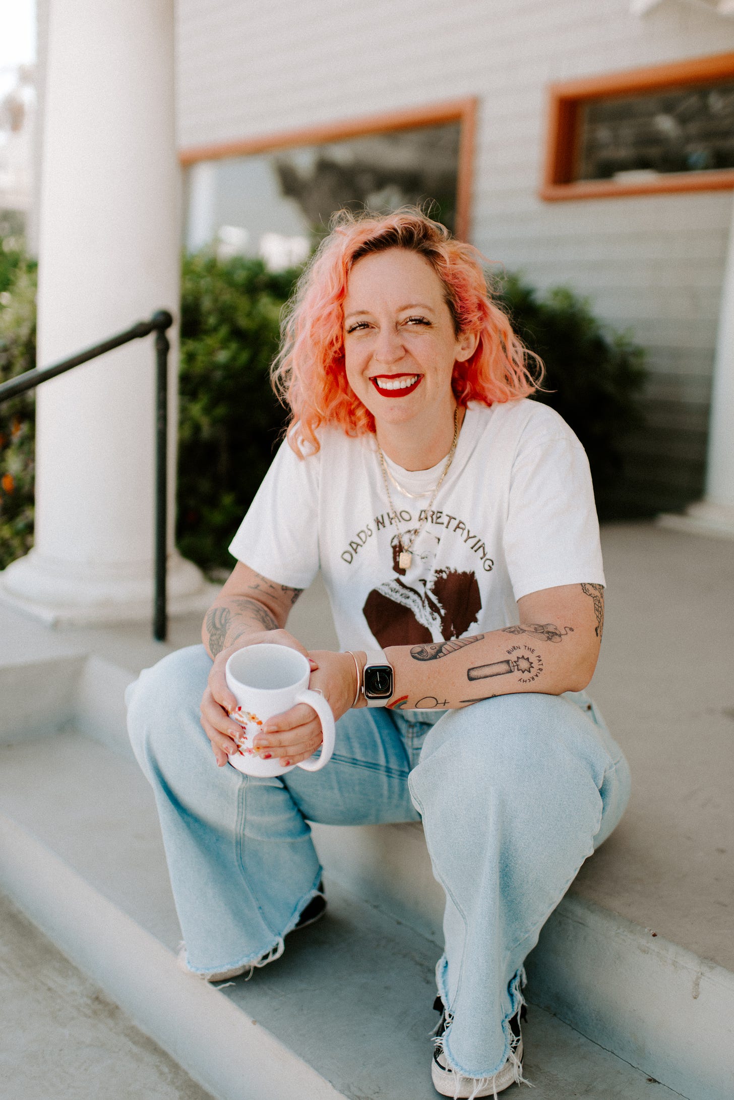 a white woman with pink curly hair smiles at the camera. She has red lipstick on and is sitting on a porch in a white tshirt and jeans holding a mug. 