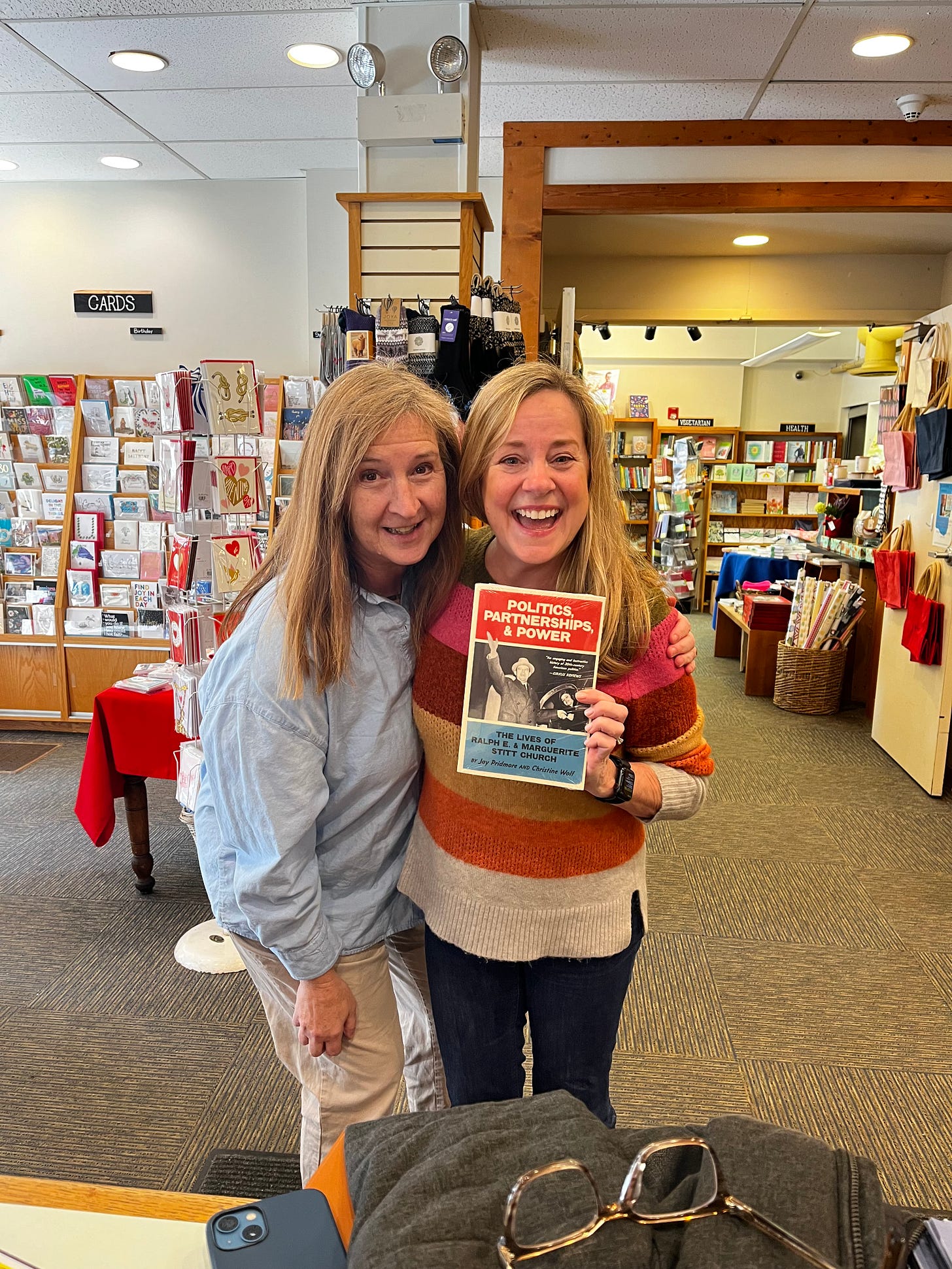 Two blonde women stand in a bookstore smiling. One woman, Memoir Coach Christine Wolf, holds her debut book in her hands for the very first time. The book is Politics, Partnerships, & Power: The Lives of Ralph E. and Marguerite Stitt Church. 