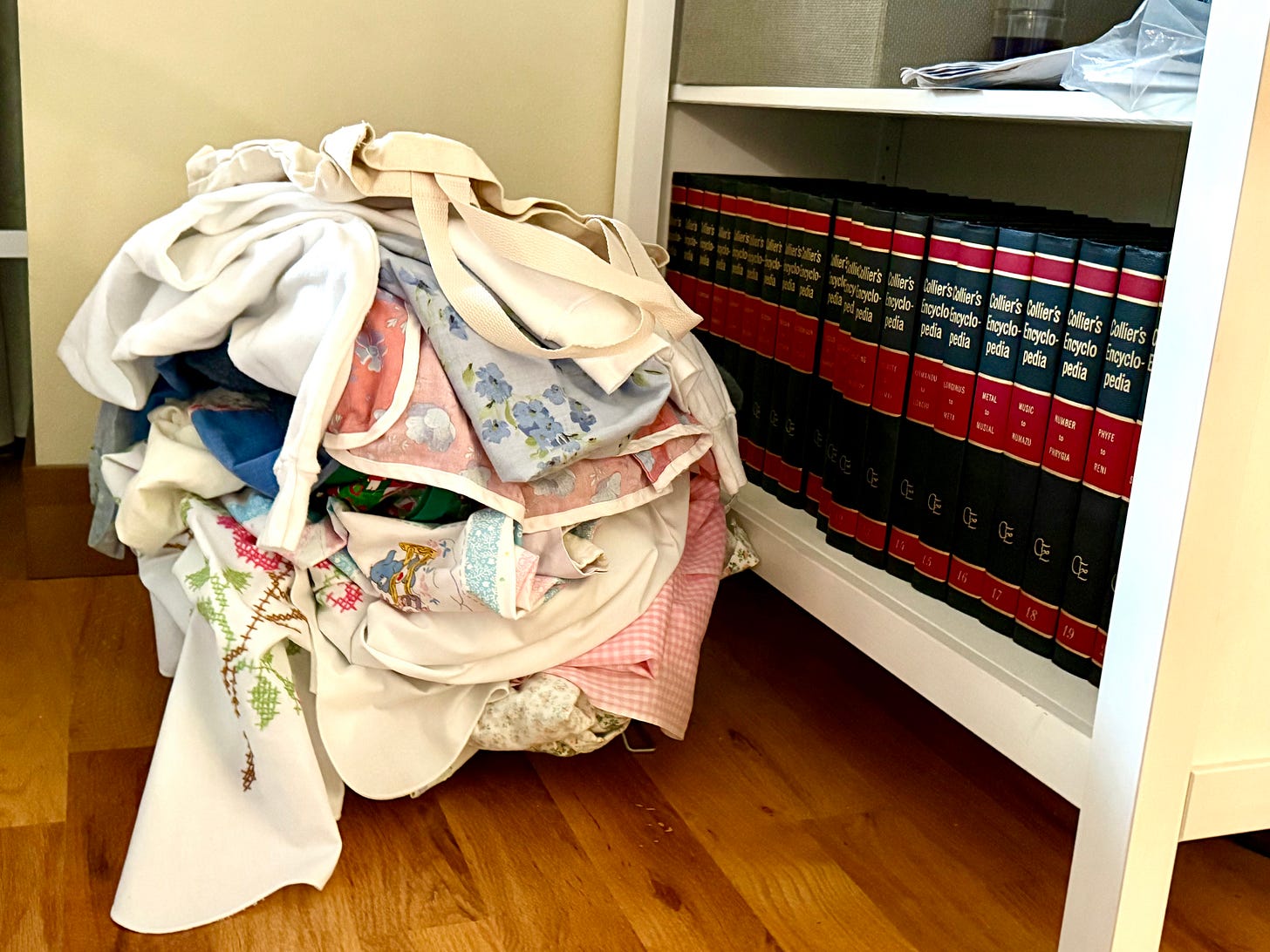 A pile of unfolded laundry with various patterns and fabrics sitting on the floor next to a bookshelf containing a set of encyclopedia volumes with red and black spines A pile of unfolded laundry with various patterns and fabrics sitting on the floor next to a bookshelf containing a set of encyclopedia volumes with red and black spines