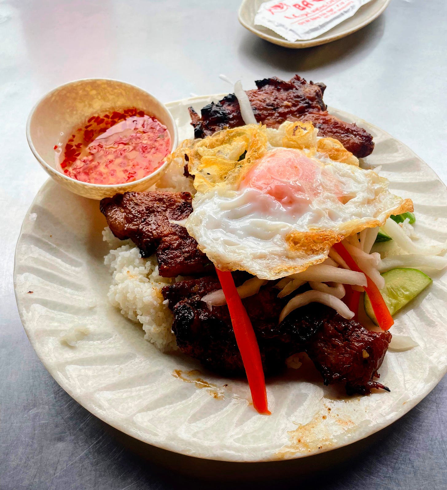 A plate of food sits on a metal table — a bed of rice, a large piece of meat topped with an egg, small julienned vegetables, and a small bowl of sauce.