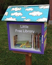 Little Free Library image with a roof showing white clouds on a blue background and a clear glass front showing the books inside.