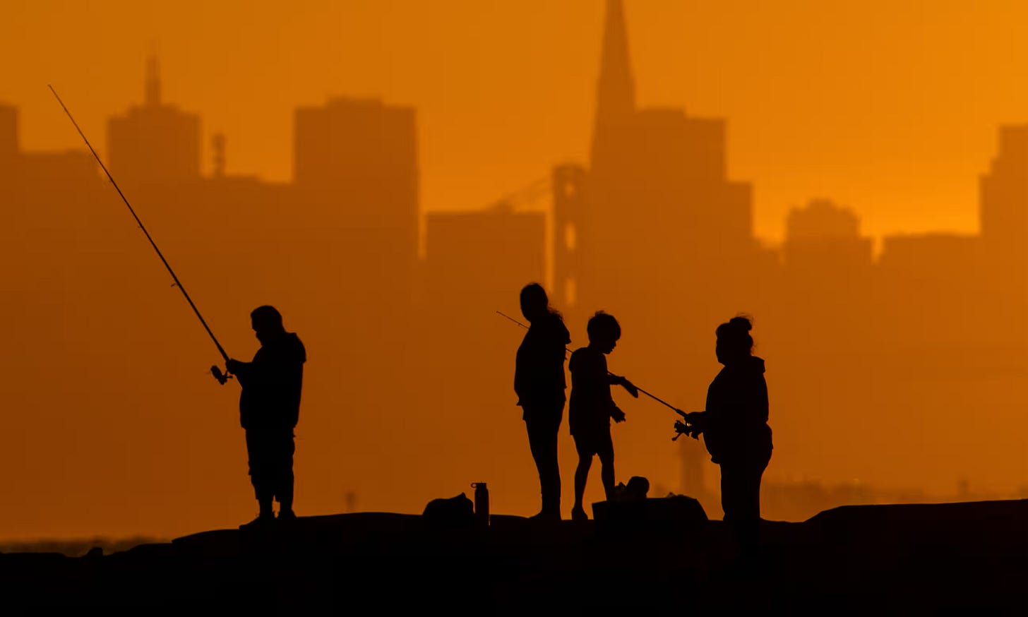 In the foreground of a photograph, silhouettes of people can be seen fishing. In the background is the skyline of a city shrouded in orange haze.