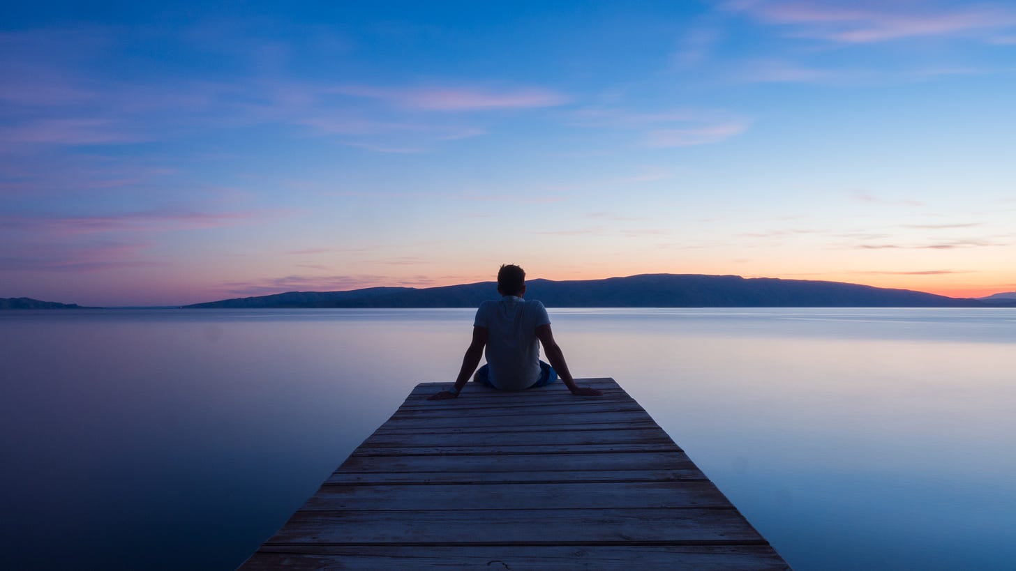 A man sits on the edge of a wooden pier overlooking a lake and mountains. The sunrise is pink on the horizon and dark blue overhead.