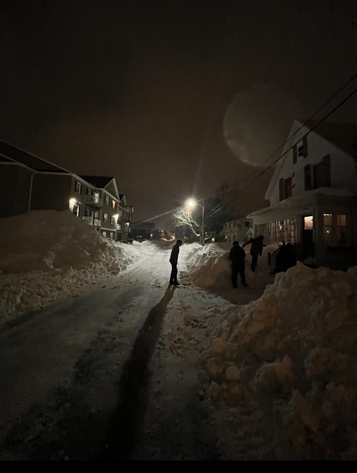 A snowy winter scene on my street. the sky is dark. we're coming home from Dairy Queen by foot. The kids are walking past giant snow drifts to get to our house.