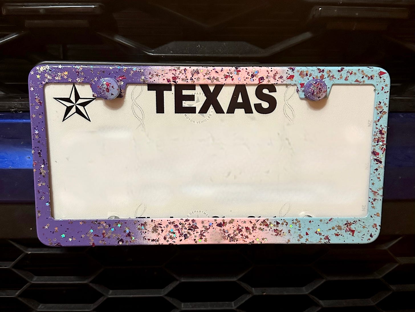 A photo of Shohreh's license plate holder which is spray painted in an ombre pattern of purple, pink, and blue with multi-color glitter flakes overlayed over it
