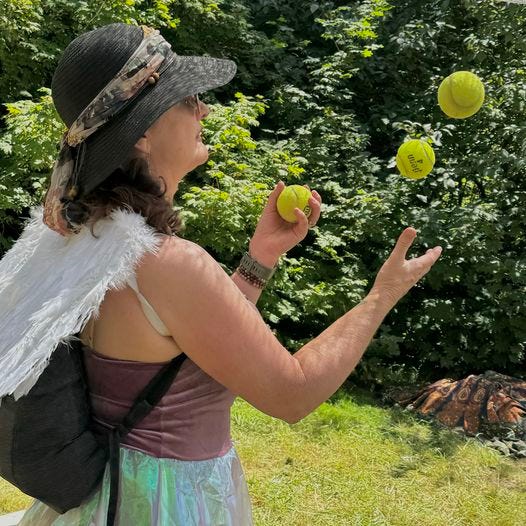 Woman in black hat and white feathered wings and ballet dress juggling balls in park surrounded by green bushes.