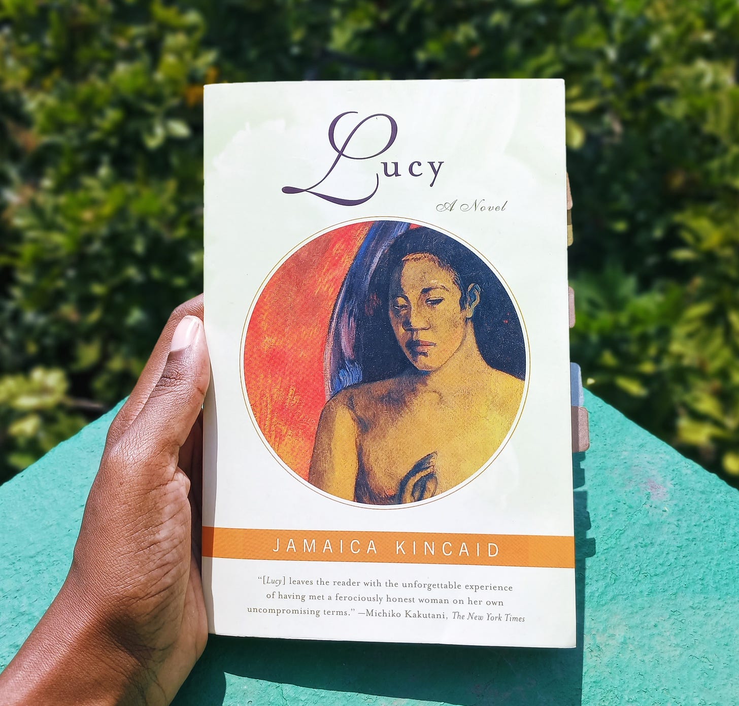 My black hand holds a paperback copy of Lucy on a sea green wall with trees below. Cover design is a pale green watercolour backgroud with a detail of Paul Gauguin's "Savage Poems": an oval frame, deep orange background, brown female figure with long black hair holding one hand in front of her chest, eyes downcast in a sidelong glance, a curve of her purple wings behind her.
