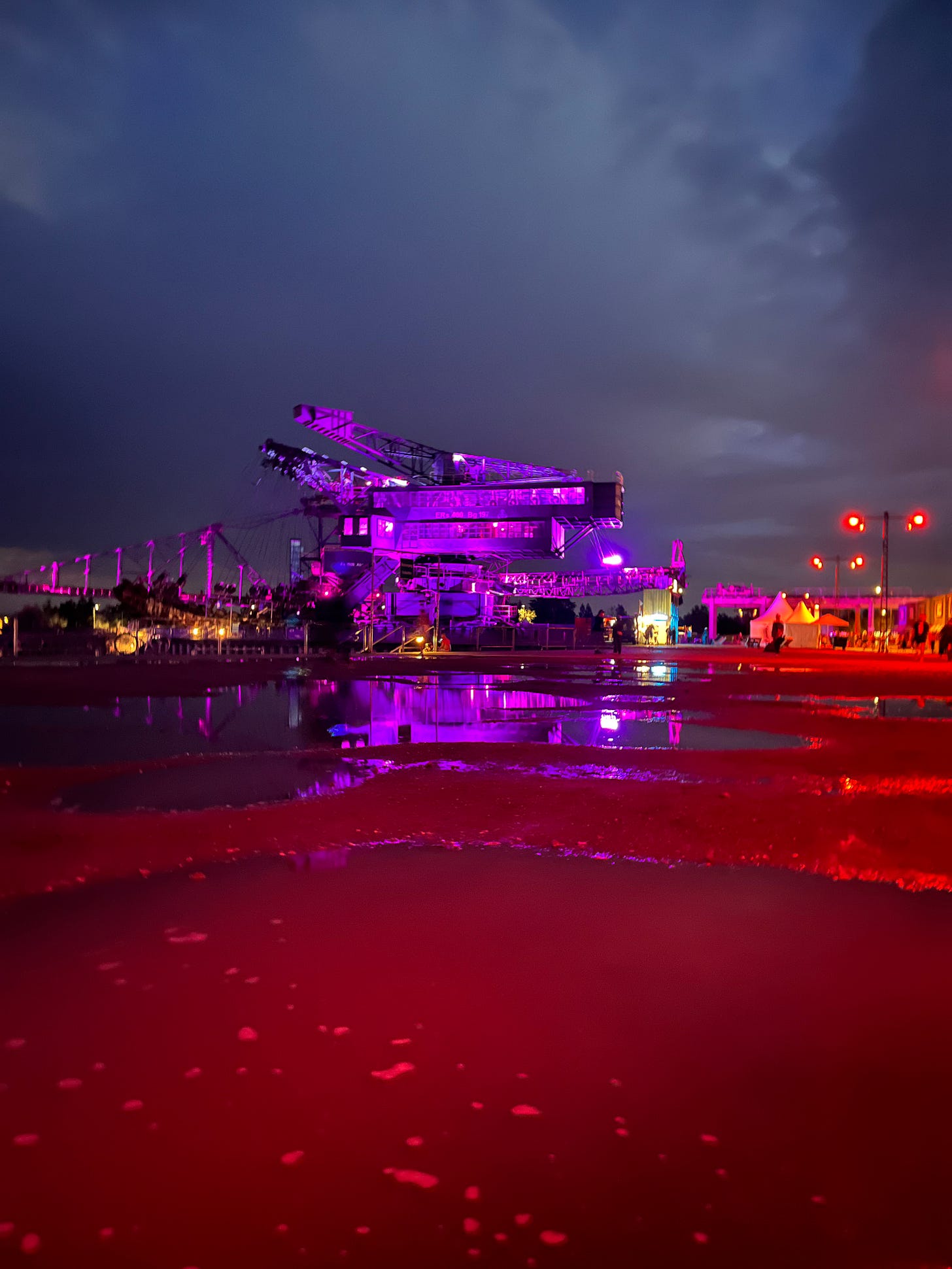 huge mining crane at night, lit up in purple. the light is reflected in the puddles on the ground at whole festival.