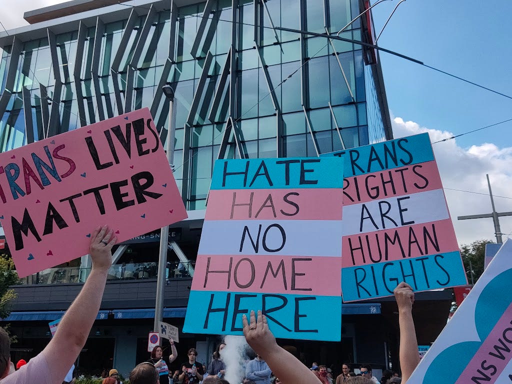 signs reading "trans lives matter" "hate has no home here" and "trans rights are human rights"