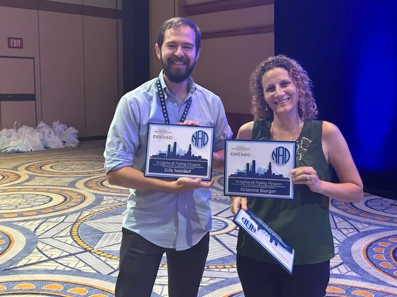 Erik Nordlof (bearded, in a button-up blue shirt) and Brianne Burger (curly-haired, in a green sleeveless top) stand together on a hotel ballroom floor, holding up their award certificates from the NAD. Both are smiling.