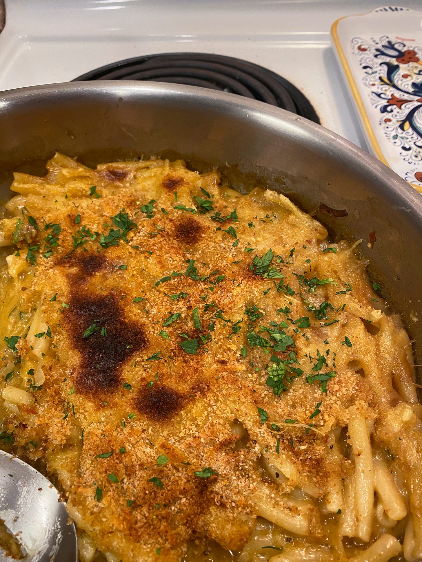 A stainless steel pan of mac & cheeze, with browned breadcrumbs and chopped parsley on top. A serving spoon is just visible in the lower left corner of the frame.