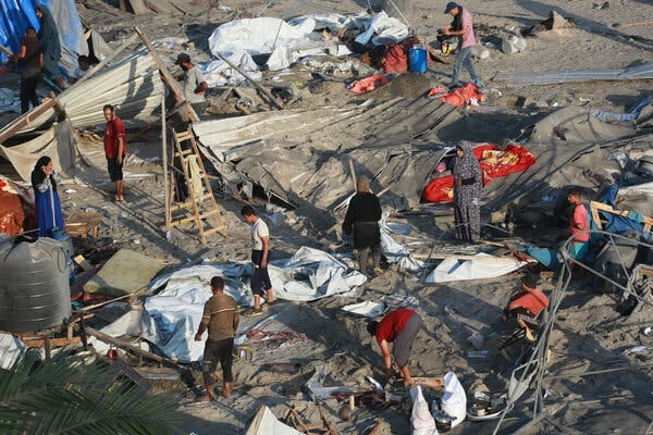 A group of men and women look at badly shredded white tents in a sandy field. 