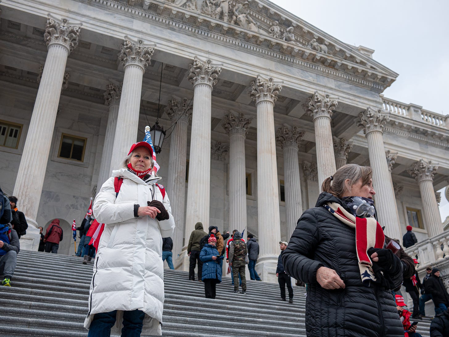 A woman in a long white down coat and red "Make America Great Again" hat stands on the steps of the US Capitol on Jan 6, 2021. A red "Trump" mask hangs from her chin and a US flag attached to a short flag pole rests on her left shoulder. She is older, with grey hair, and she looks off into the distance as others are randomly gathered on the steps.