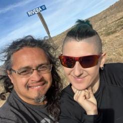 Photo of Lyric and David Rivera, under Rivera RD at sign in the NM dessert. David is a long haired brown skin man with a teethy smirk, and Lyric is a fair skin creature with short green hair and shaved sides.