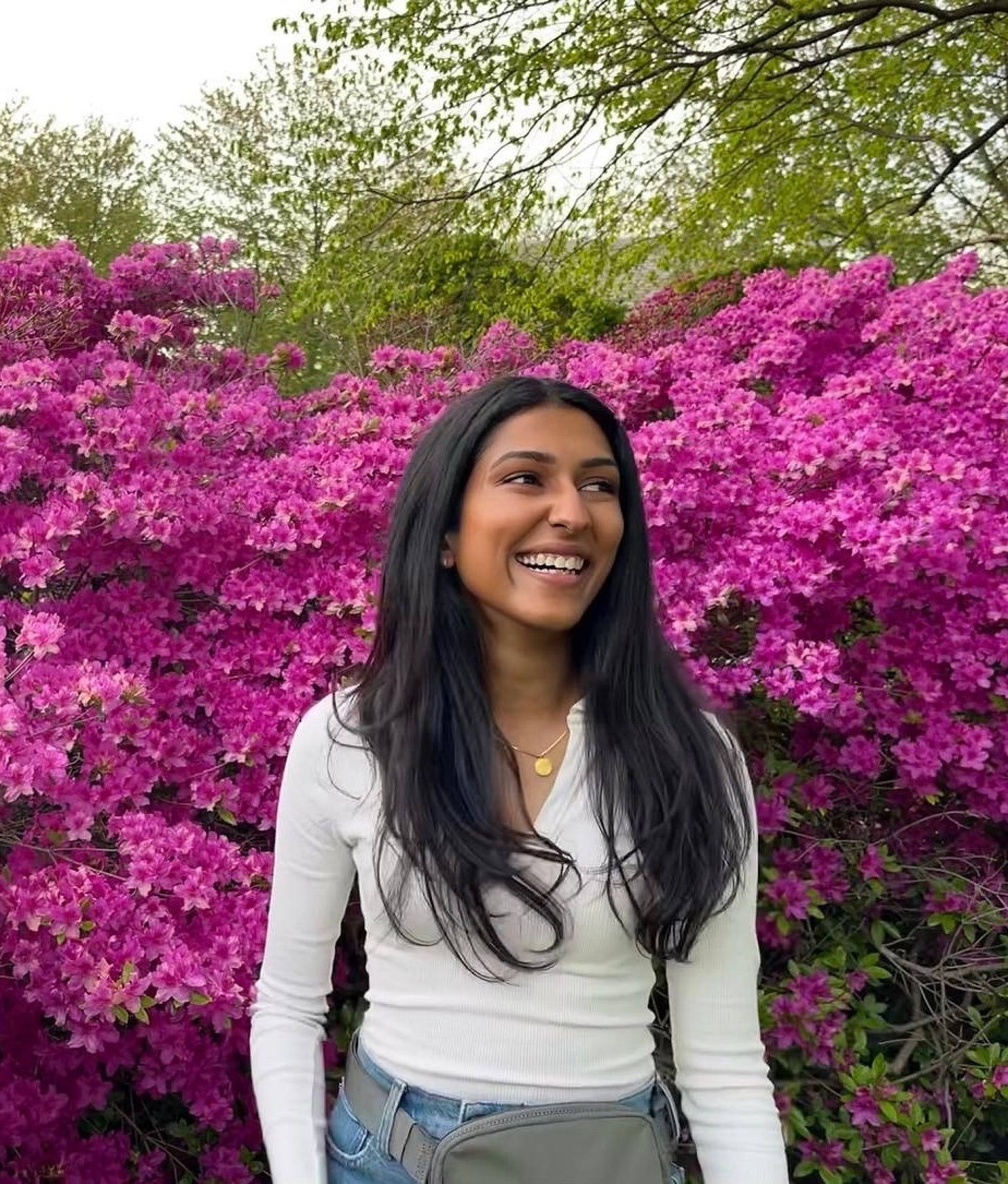 Abi smiling in a white top, blue jeans, and bag looking away from the camera in front of a wall of pinkish purple flowers.