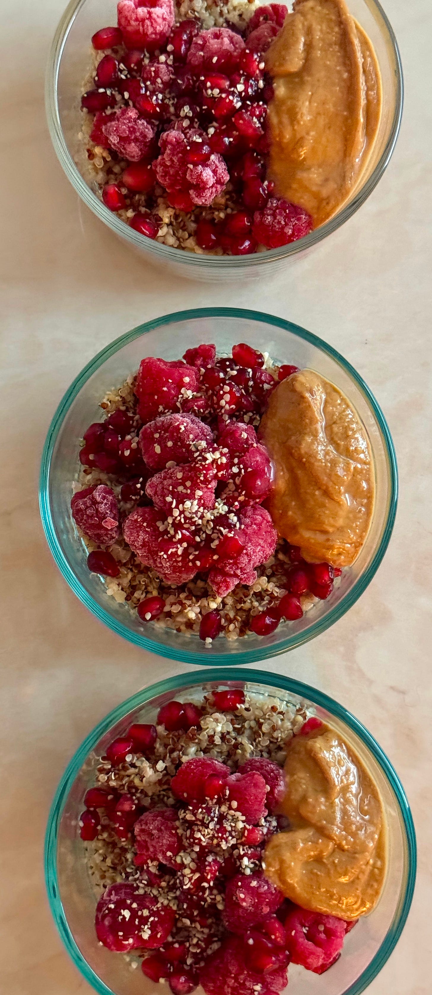 A bright raspberry quinoa breakfast bowl topped with fresh raspberries, pomegranates, hemp seeds, and a drizzle of nut butter in a clear bowl on a light counter.