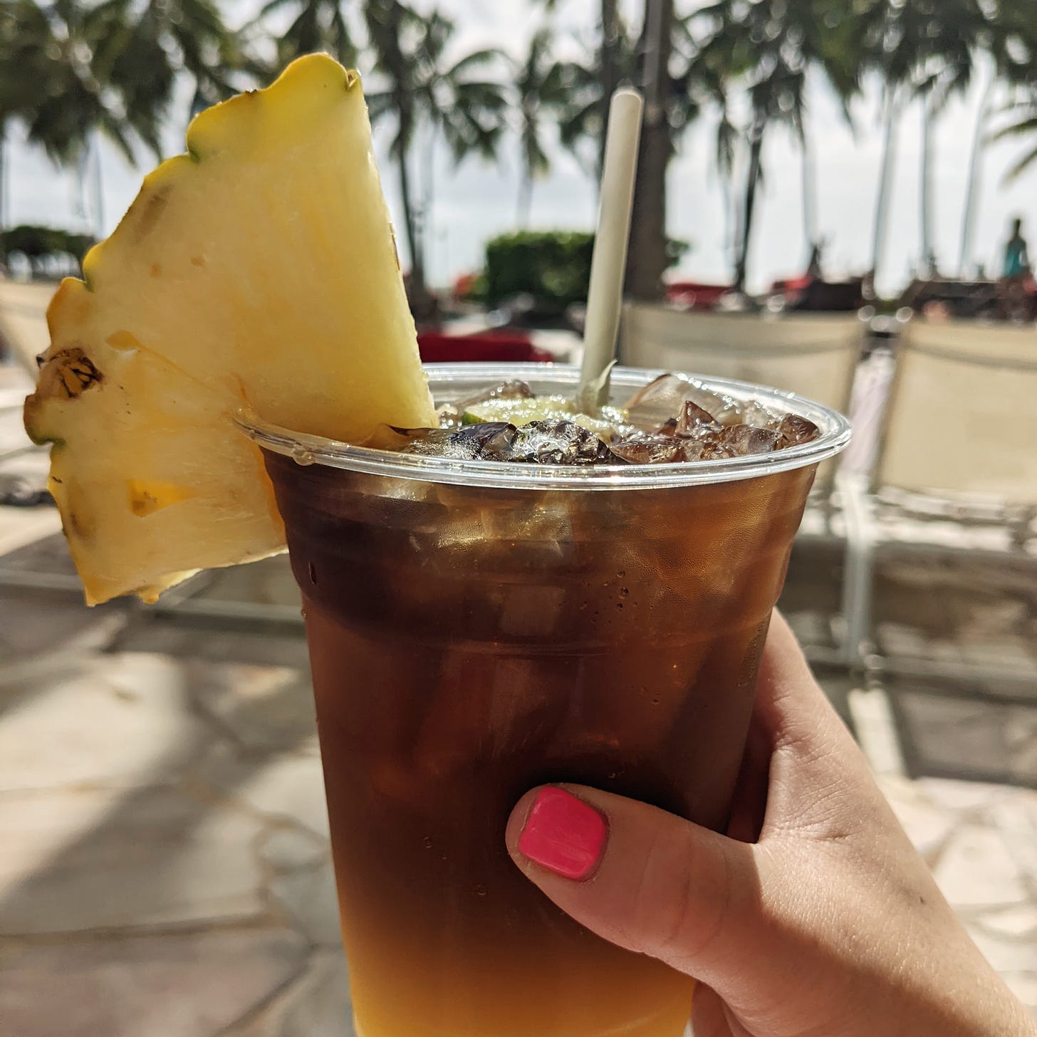 A woman's hand holding a plastic cup with a mai tai cocktail and a pineapple wedge
