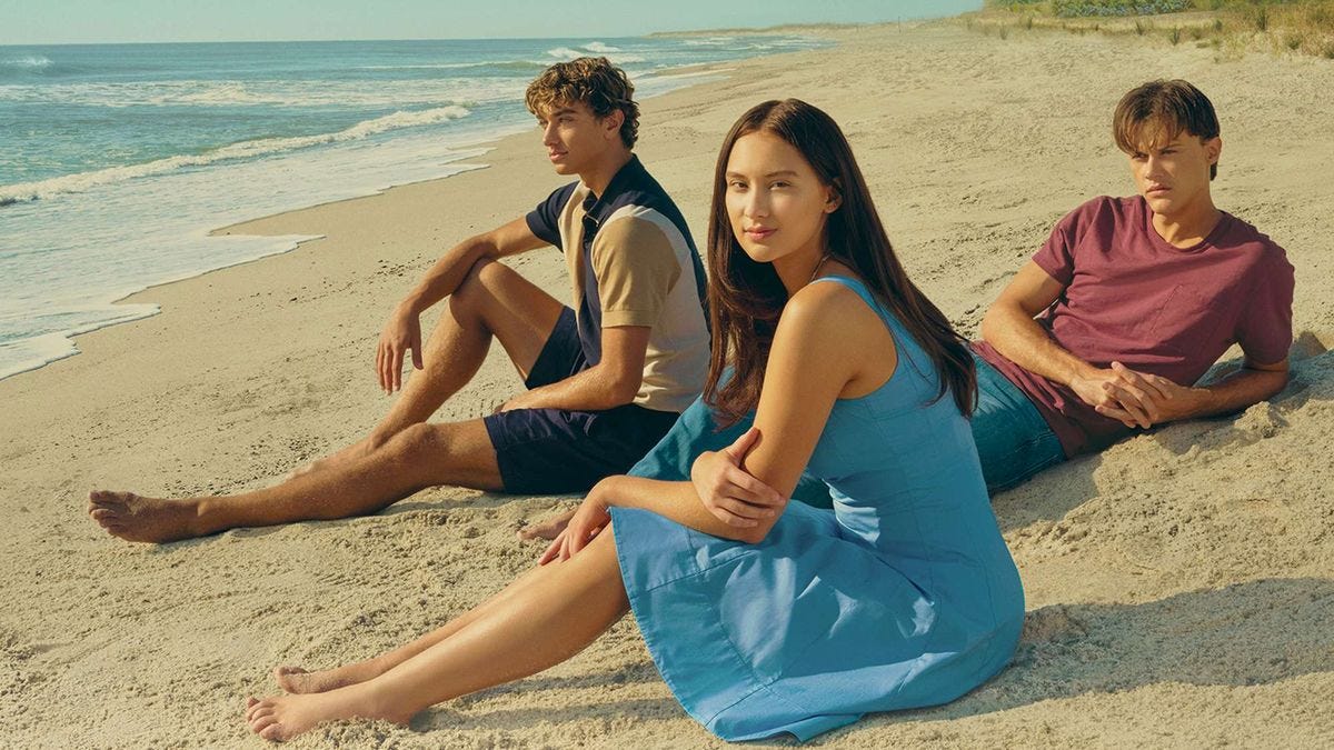 Three characters (two boys and a girl in a blue dress) sitting at the beach, from the show "The Summer I Turned Pretty."