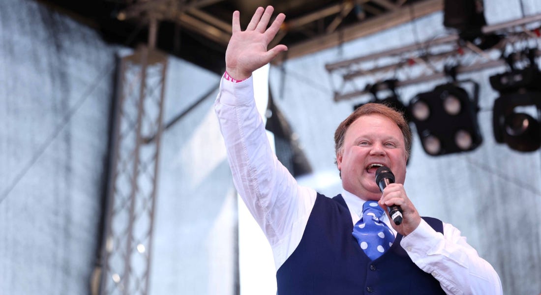 Justin Fletcher wearing a blue waistcoat and shirt holding a microphone waving to the crowd Justin Fletcher wearing a blue waistcoat and shirt holding a microphone waving to the crowd