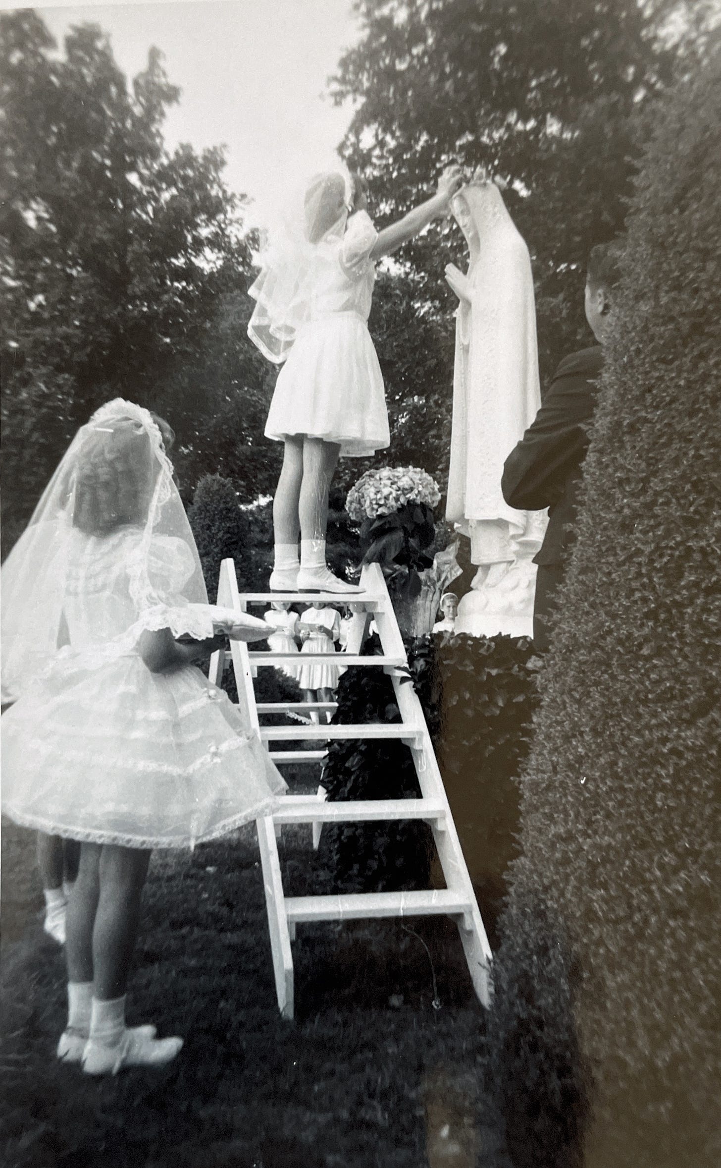 A seven year girl dressed in a white first communion dress and veil, standing on a ladder crowning the May Queen, with another girl similarly dressed holds a pillow for the crown 