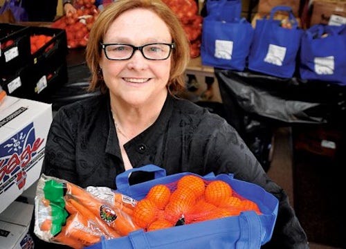 Photograph of a smiling woman, in a church hall filled with boxes of food, holding a large bag in which can be seen carrots, oranges and other food.