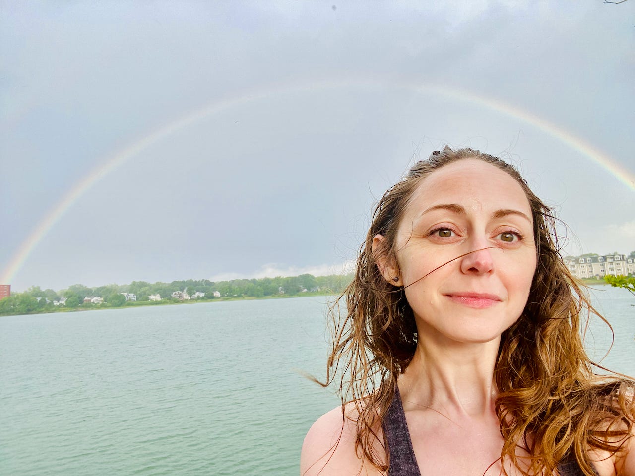 Self-portrait of Kate standing in front of a rainbow against a cloudy sky. Kate’s hair and skin are wet from running out into the rain, and she wears an expression of bewildered gratitude.