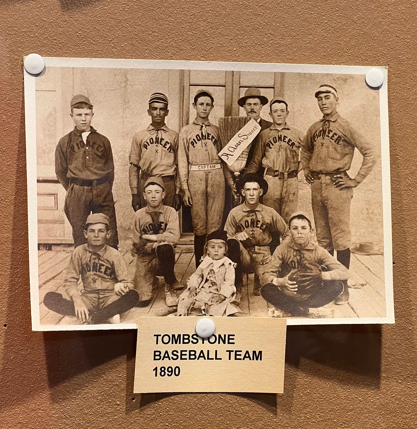 an image of a photograph on a bulletin board of the tombstone pioneers baseball team. it's mostly teenage kids with one toddler in the front, and one is holding a broom that says 'a clean sweep' on it.