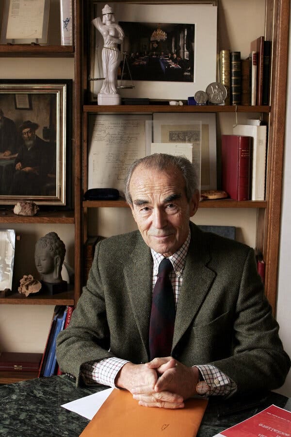 A portrait of Robert Badinter, wearing a jacket over a shirt and tie while sitting in front of his desk at home.