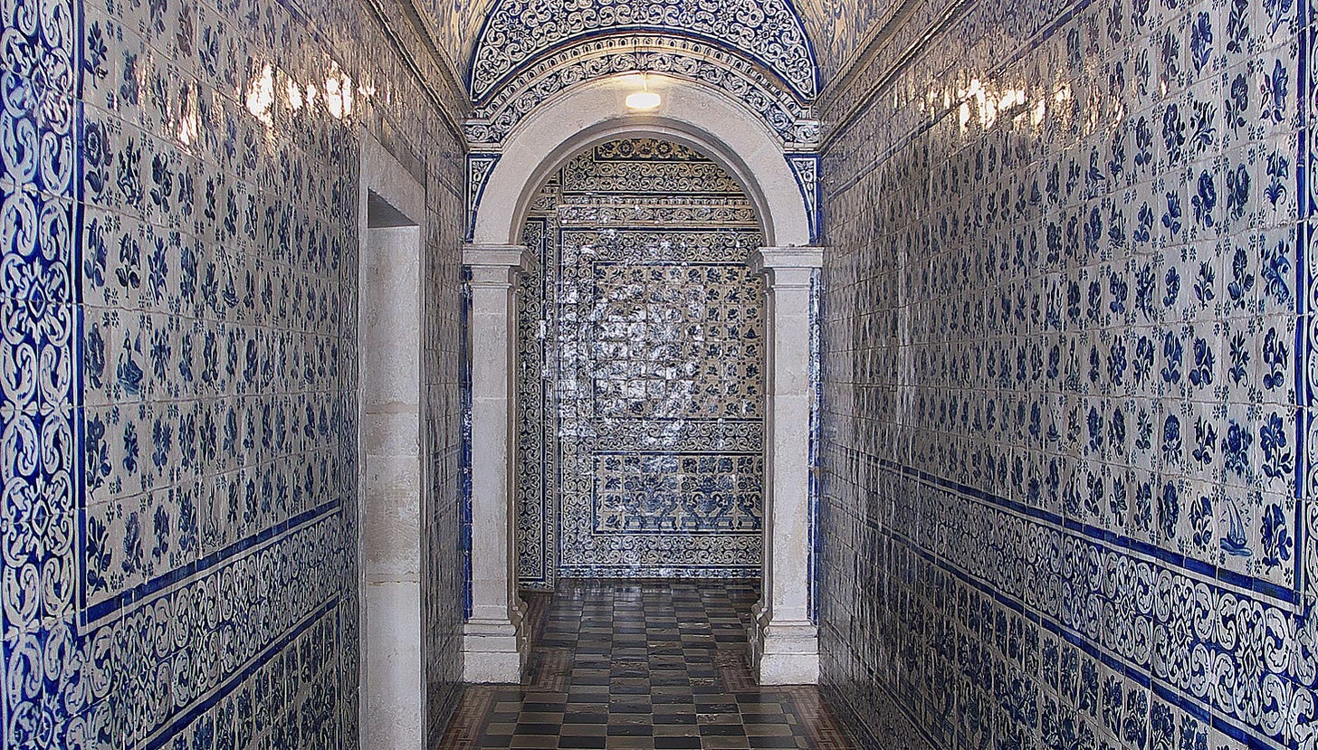 A white archway at the end of a corridor is surrounded on all sizes by busy patterns on traditional blue and white tiles. The floor is black and brown chec