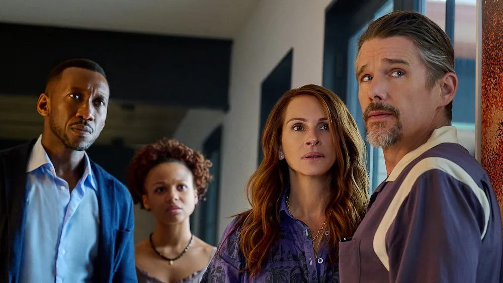 A still from the film Leave The World Behind, four people stand inside a home, with white walls and wood visible behind them. A slim Black man in a blue button-up shirt and a navy cardigan looks concerned, next to his daughter who has short, curly hair, a necklace, and an off-the-shoulder dress. Next to her is a white woman with long hair and a peasant-style blouse, and next to her is her husband, a white man with a salt and pepper goatee and short hair in a white and purple hoodie.