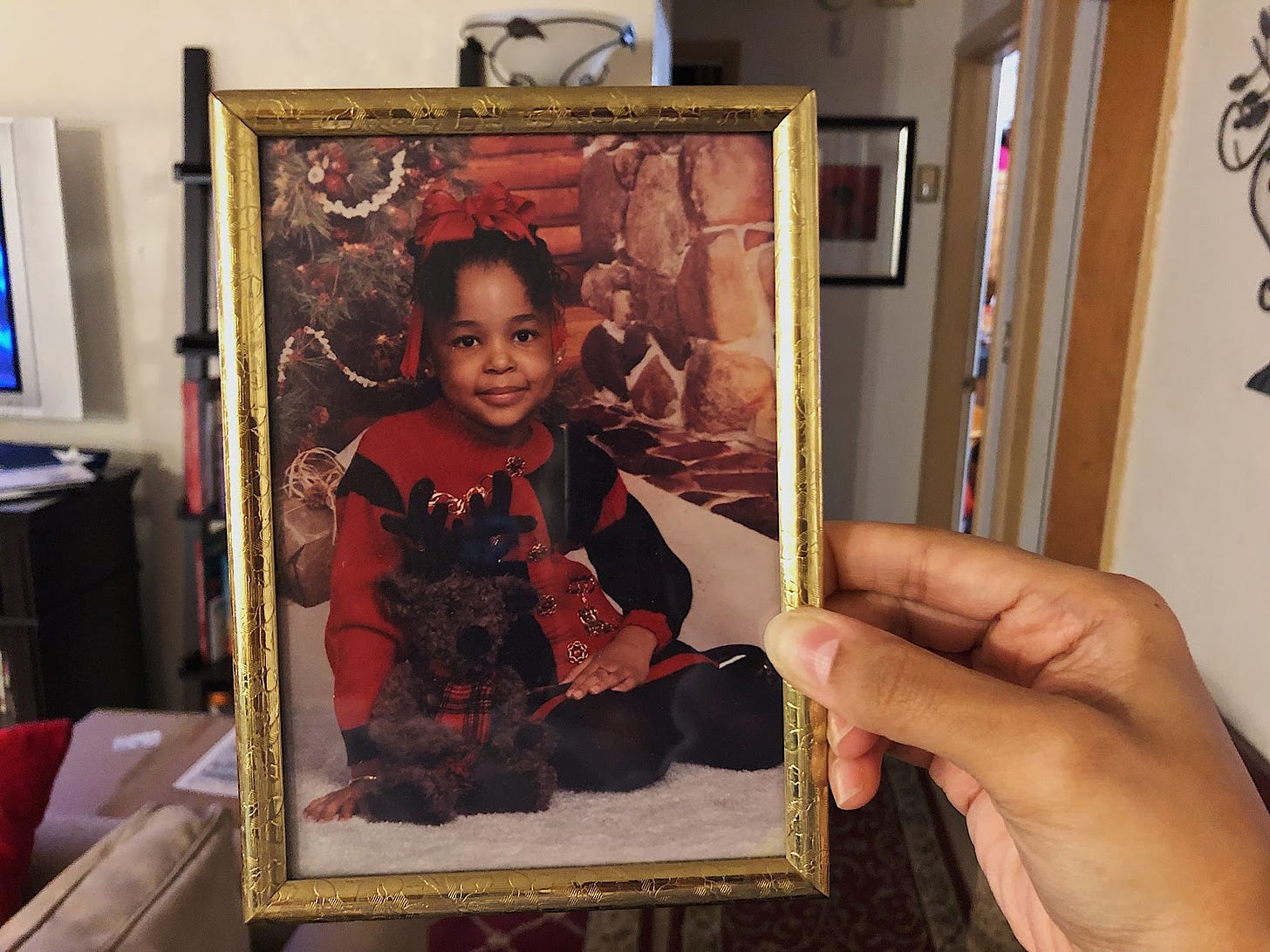 A hand holding up a framed picture of a black girl in red and black Christmas clothes and a stuffed reindeer posing for a photo.
