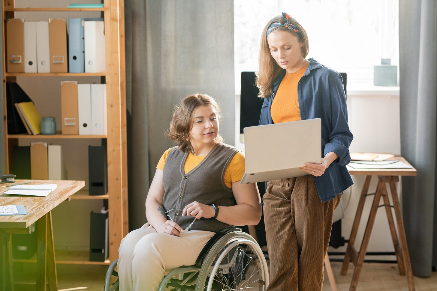 Two women are looking at a laptop in an office. One woman is wearing business casual clothes and is sitting in a wheelchair. The other woman is similarly dressed and is holding the laptop.