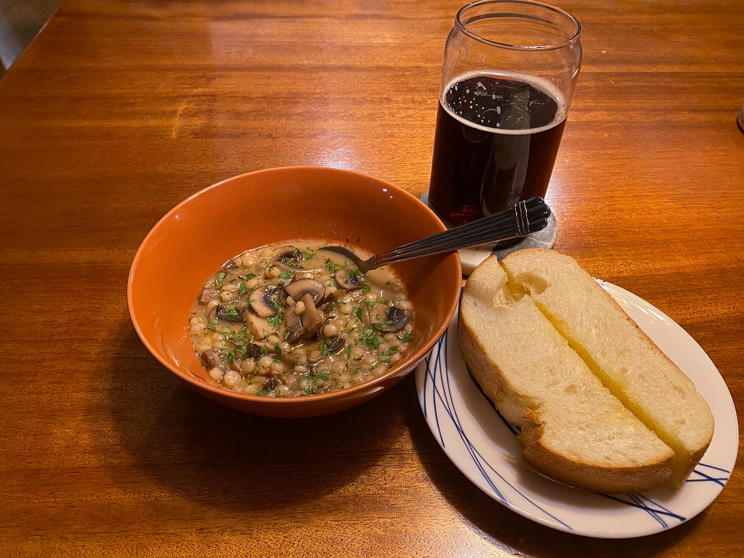 An orange bowl of the soup described above, with parsley sprinkled over the top and a spoon resting at the edge. Next to it is a white and blue plate with a slice of garlic bread on it, cut in half crosswise. Above it is a mostly-full glass of dark beer.