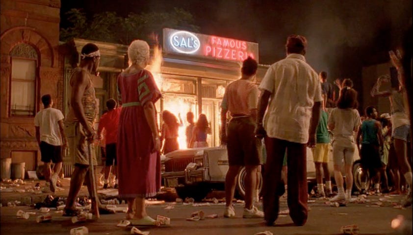 This image shows several spectators standing on the street and facing Sal's Famous Pizzeria, which is on fire, in the famous concluding sequence of Spike Lee's 1989 film Do the Right Thing.