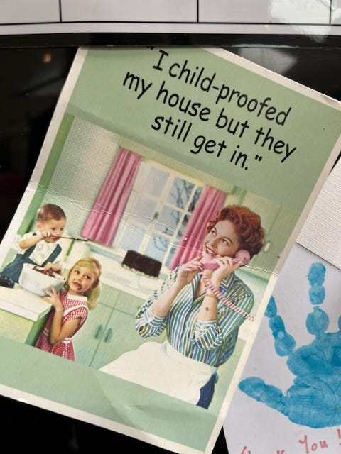 Old-fashioned postcard on the fridge of a woman talking on her phone in the kitchen. Text reads "I child-proofed my house but they still get in." There are kids covered in chocolate cake batter behind her.
