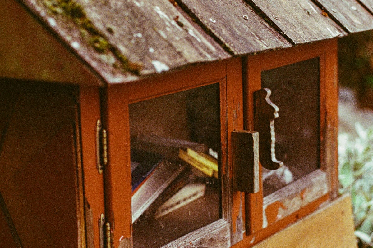 A small wooden chest with hinged, windowed doors and a mossy tiled roof serves as a library for used books, stacked inside against each other.