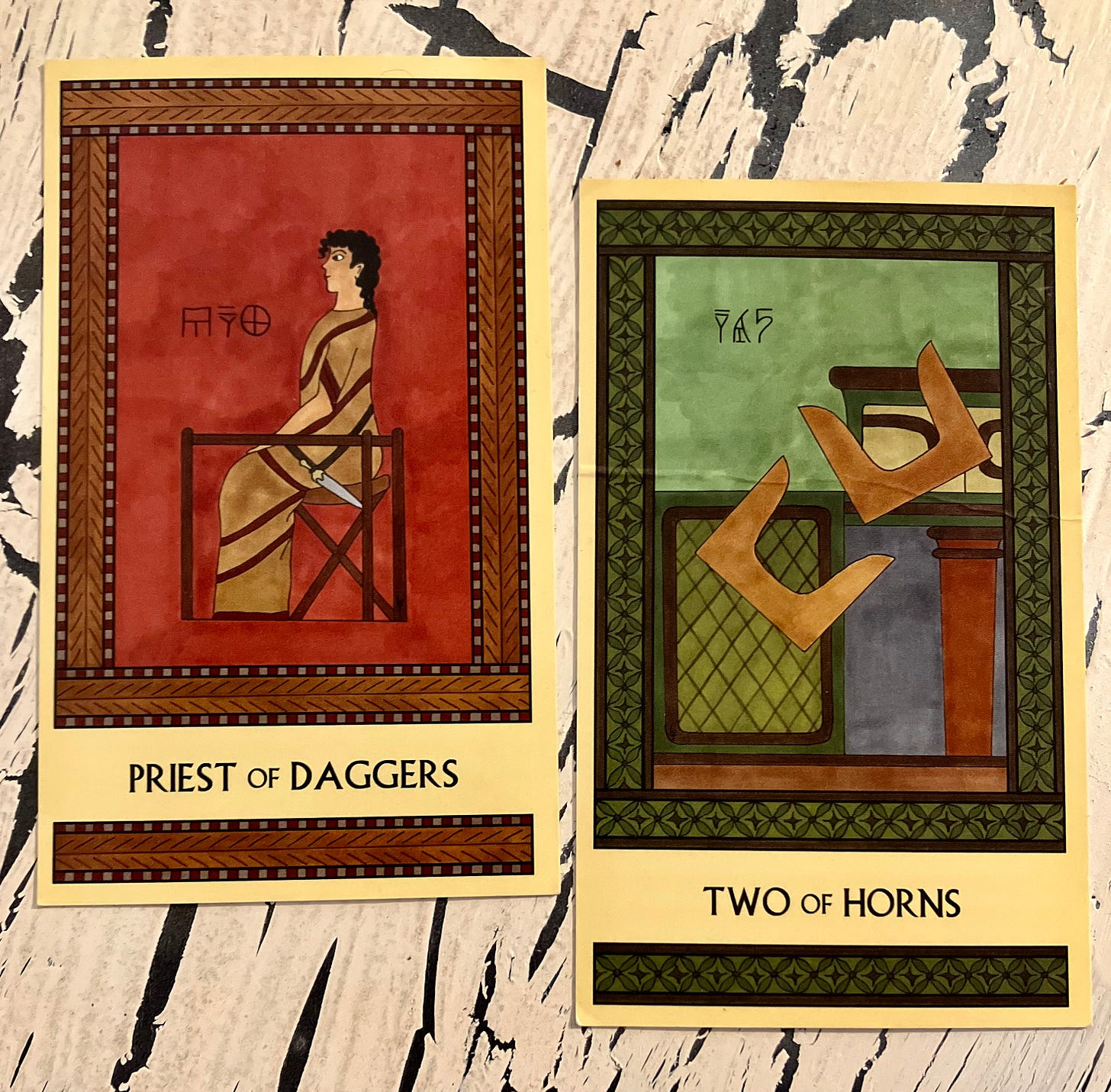 Two Minoan Tarot cards on a crackled cream colored surface. The Priest of Daggers is in shades of deep ted and brown. It shows a Minoan man in a long robe, seated facing left, with a dagger at his waist. The Two of Horns is in shades of green and tan. It shows two pairs of sacred horns tumbling off a shrine.