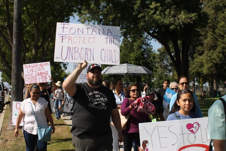 People marching in protest of Planned Parenthood holding signs. One sign reads Fontana, Protect the Unborn Child