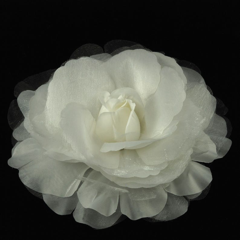 May include: A large white fabric flower with a ruffled edge. The flower is made of multiple layers of sheer fabric and has a delicate, feminine appearance.