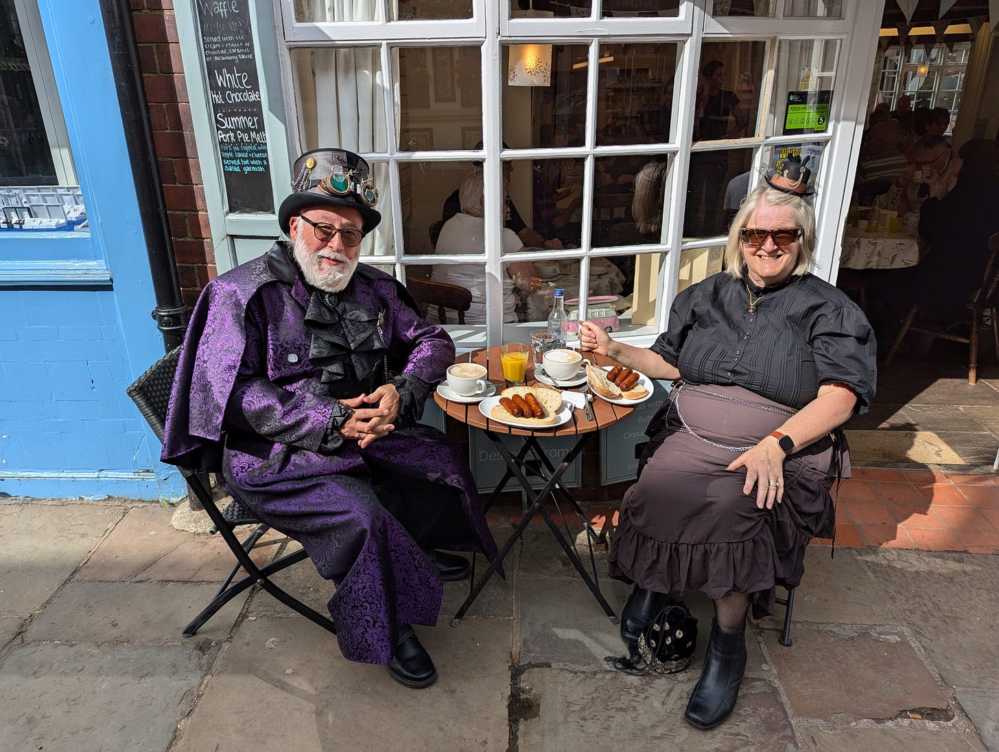 A steampunk couple smile into the camera, sat outside a cafe with their breakfast on the table between them