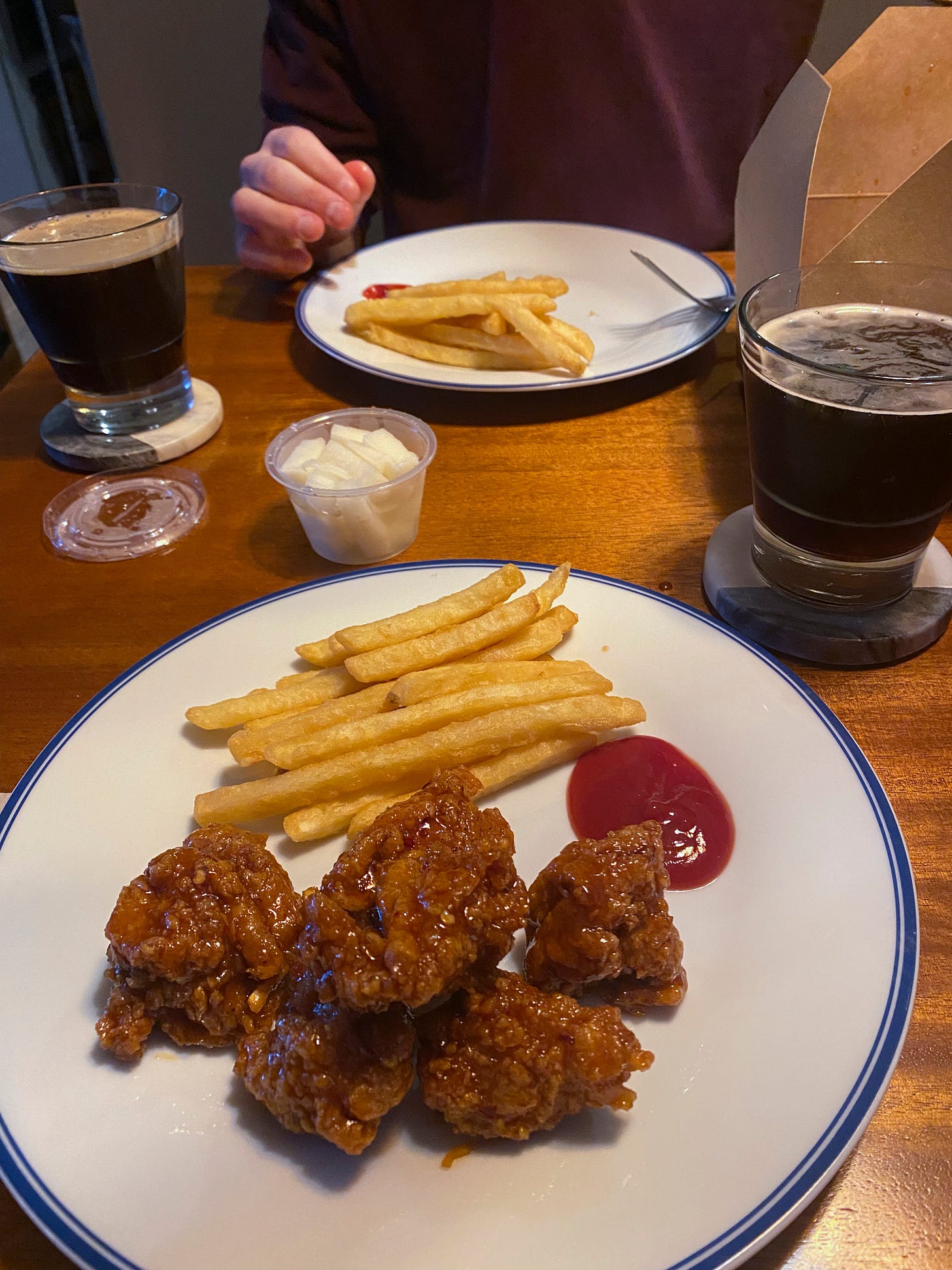 Two plates with pieces of fried chicken in a spicy-sweet sauce, thin and crispy fries, and a little pool of ketchup. Between the plates is a plastic container of pickled daikon, and two glasses of dark beer sit on coasters at the edges of the plates. Just visible in the upper right of the frame is the box holding the rest of the chicken pieces.