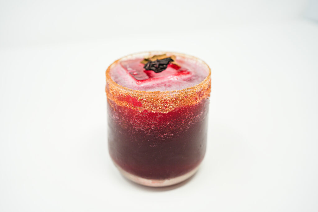 A cinnamon hibiscus mezcal margarita in a glass rimmed with cinnamon, showcasing a deep red hue from the hibiscus infusion. The drink is elegantly garnished with a cinnamon stick and a vibrant red hibiscus flower, enhancing its visual appeal.
