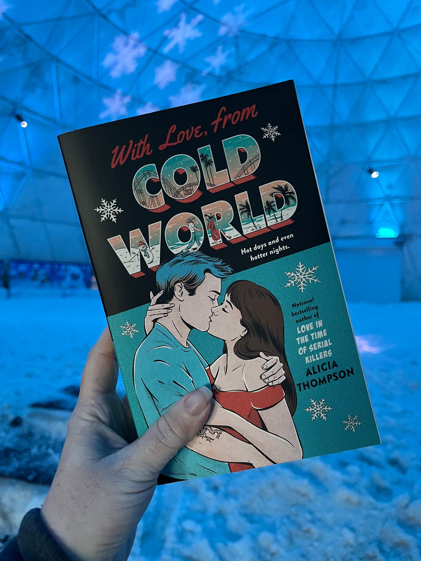 Picture of my hand holding up the teal cover copy of WITH LOVE, FROM COLD WORLD in the Igloo, which is a self-contained area with fake snow