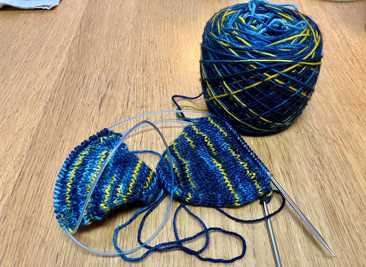 the toe-portion of two socks out of a variegated blue and yellow yarn are on a single long circular needle, sitting next to the skein they are being knit from. 