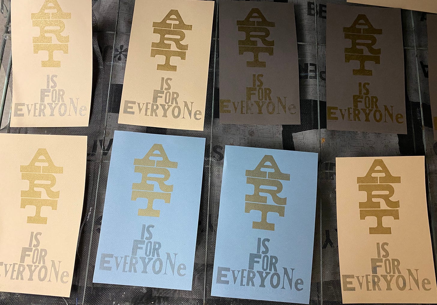 Gold and silver letterpress prints reading "Art is for everyone" on tan, blue, and grey paper, laying on a drying rack.