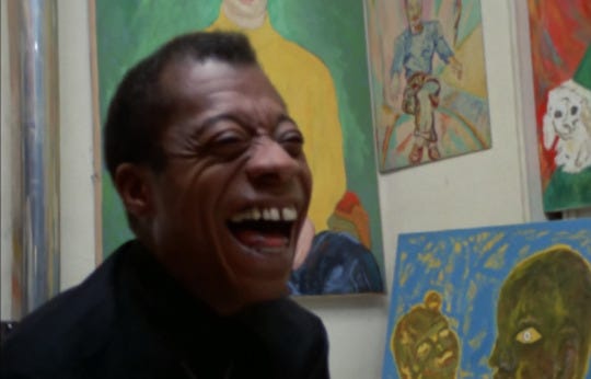 Still from a documentary showing a Black man, sitting in front of a wall of colorful paintings, laughing with a big smile as he looks to people off camera.