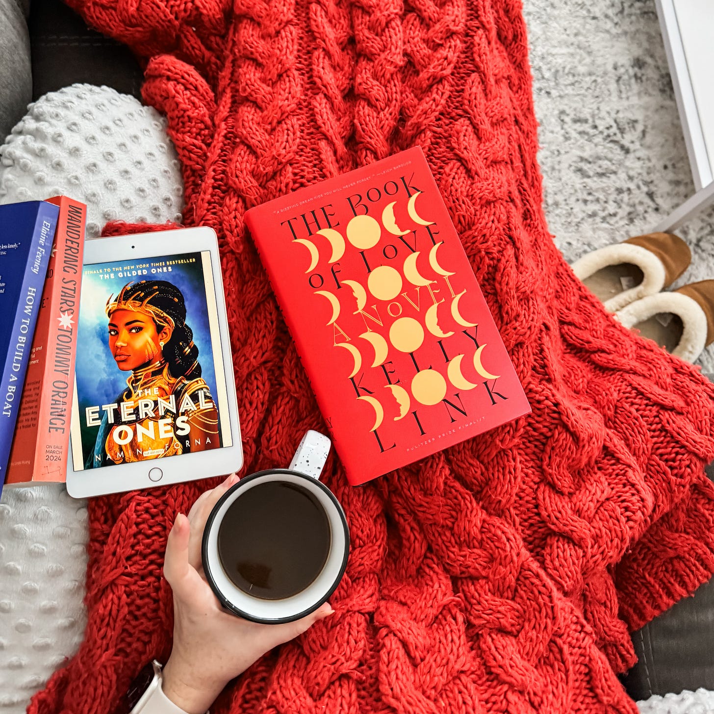 A photo of my lap, under a cozy knit blanket, including The Book of Love, three other books, and my morning coffee.