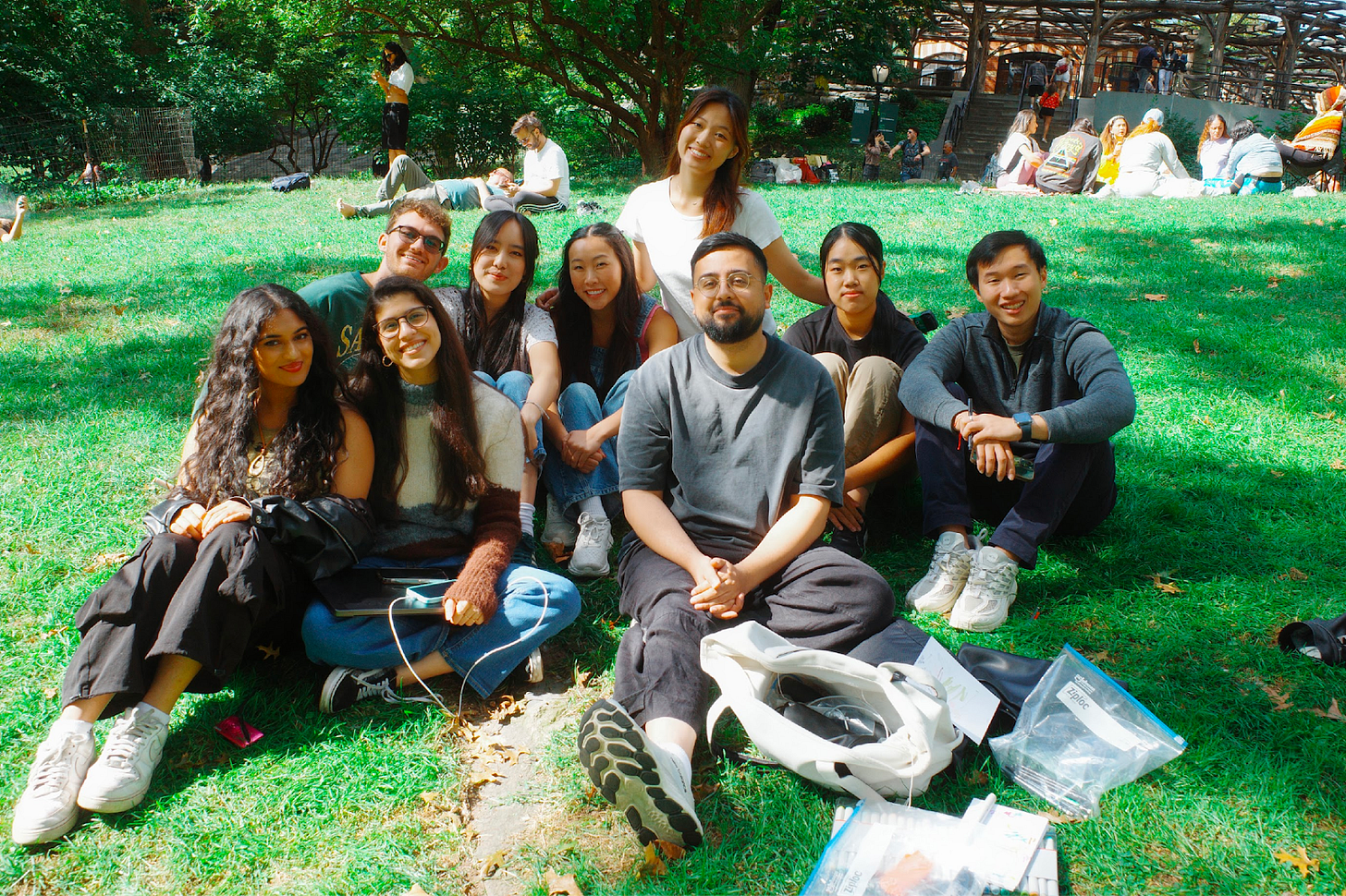 A group of nine people sitting together on grass in a park, smiling at the camera. They are casually dressed, and various bags and items are scattered in front of them. Behind them, other people are sitting and lying on the grass, enjoying the sunny day. Trees and a wooden pavilion structure are visible in the background.