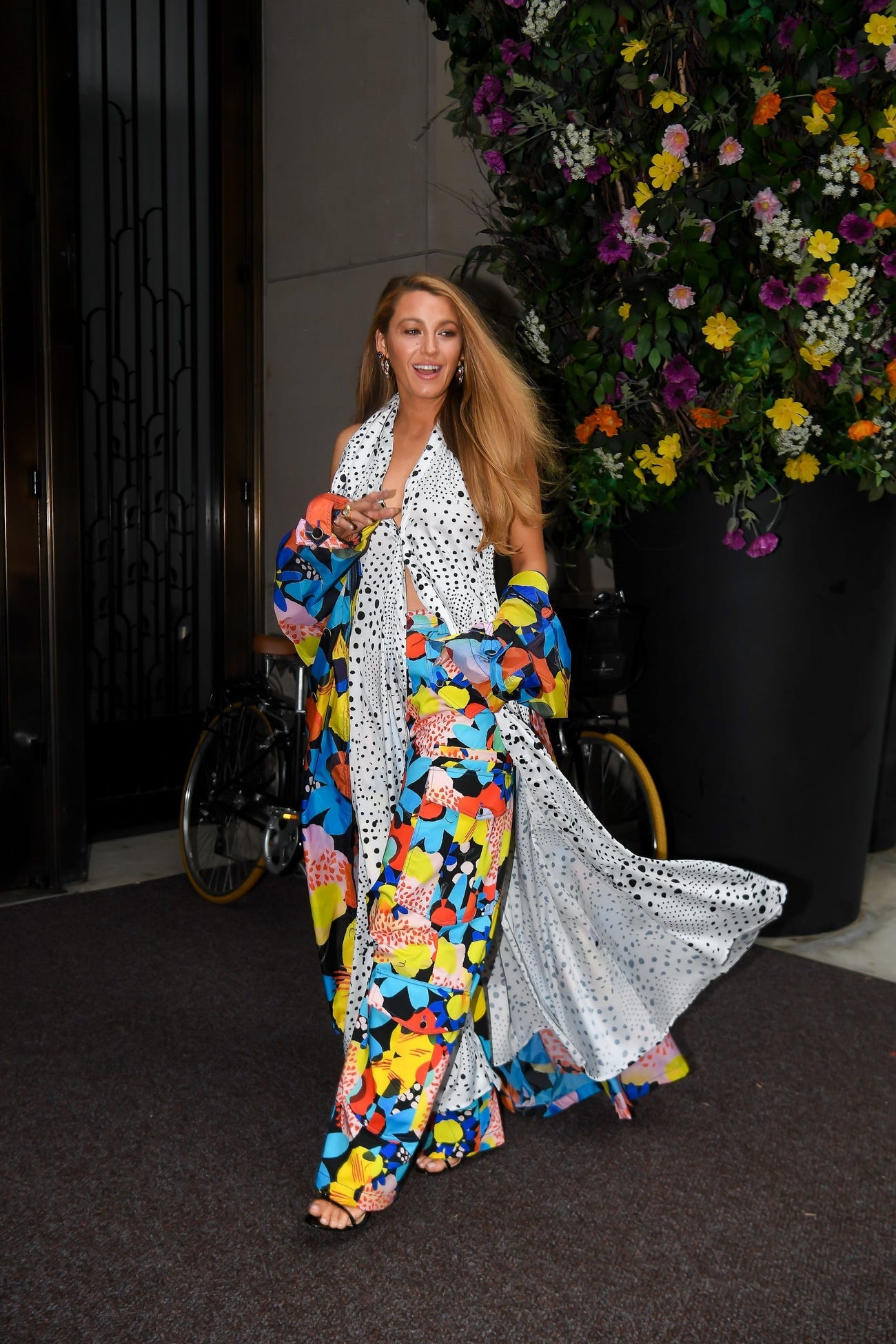 Blake Lively walks in front of a floral wall in a floral and polka-dot outfit. 